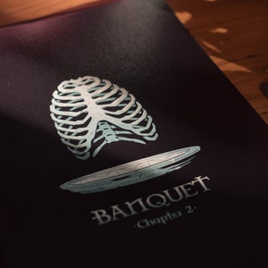 May include: A black book cover with a white ribcage and a plate illustration. The text "BANQUET - Chapter 2." is printed on the cover.
