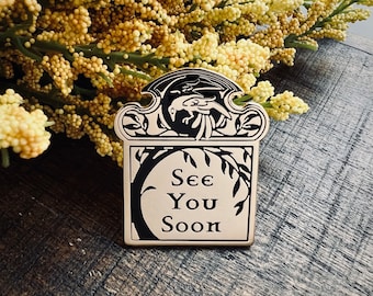 Può includere: Spilla in smalto dorata con un designo in bianco e nero di una lapide con un uccello appollaiato su un ramo. La lapide recita "See You Soon".