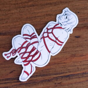 May include: A white sticker with a line drawing of a person tied up with red rope. The person has short hair and is lying on their side.