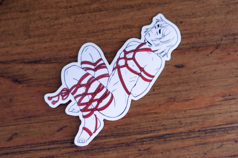 May include: A white sticker with a line drawing of a person tied up with red rope. The person has short hair and is lying on their side.