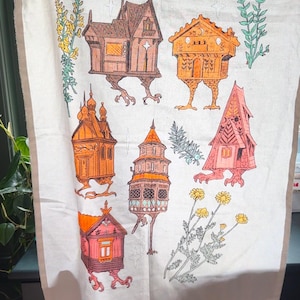 May include: A white tea towel with a colorful illustration of six houses with legs. The houses are in various styles and colors, including red, orange, brown, and pink. The houses are surrounded by green and yellow flowers.