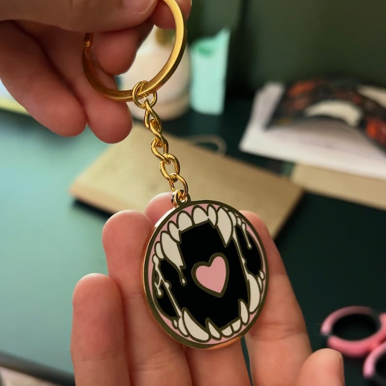 May include: Gold keychain with a round enamel charm featuring a black and white illustration of a mouth with fangs and a pink heart inside.