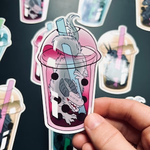 Op de afbeelding: Een sticker met een grijs en wit wezen met een tandenrij, ondergedompeld in een roze en witte bubble tea met zwarte boba-parels. Het wezen houdt een blauwe rietje vast.