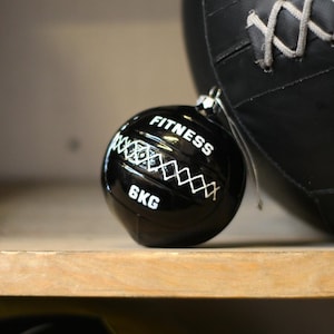 May include: A black fitness ball with white text that reads "FITNESS" and "6KG". The ball is hanging in front of a larger black exercise ball. The smaller ball has white stitching and is on a wooden shelf.