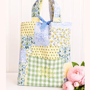 May include: A quilted tote bag featuring a patchwork design of yellow, blue, and white floral and gingham fabrics. The bag includes a green and white gingham front pocket embroidered with the yellow monogram GMM and a blue gingham ribbon tie.