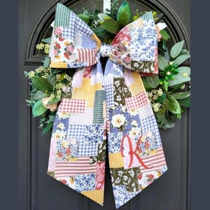 May include: A large decorative bow made of patchwork fabric featuring various floral, gingham, and striped patterns in shades of blue, yellow, pink, and green. The right tail of the bow is embroidered with a red cursive letter K. The bow is attached to a wreath.
