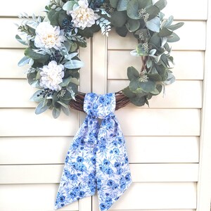 May include: A circular wreath constructed from brown twigs, adorned with green eucalyptus leaves, dusty miller foliage, and white dahlia flowers. A long fabric ribbon featuring a blue and white floral pattern is tied at the bottom of the wreath.