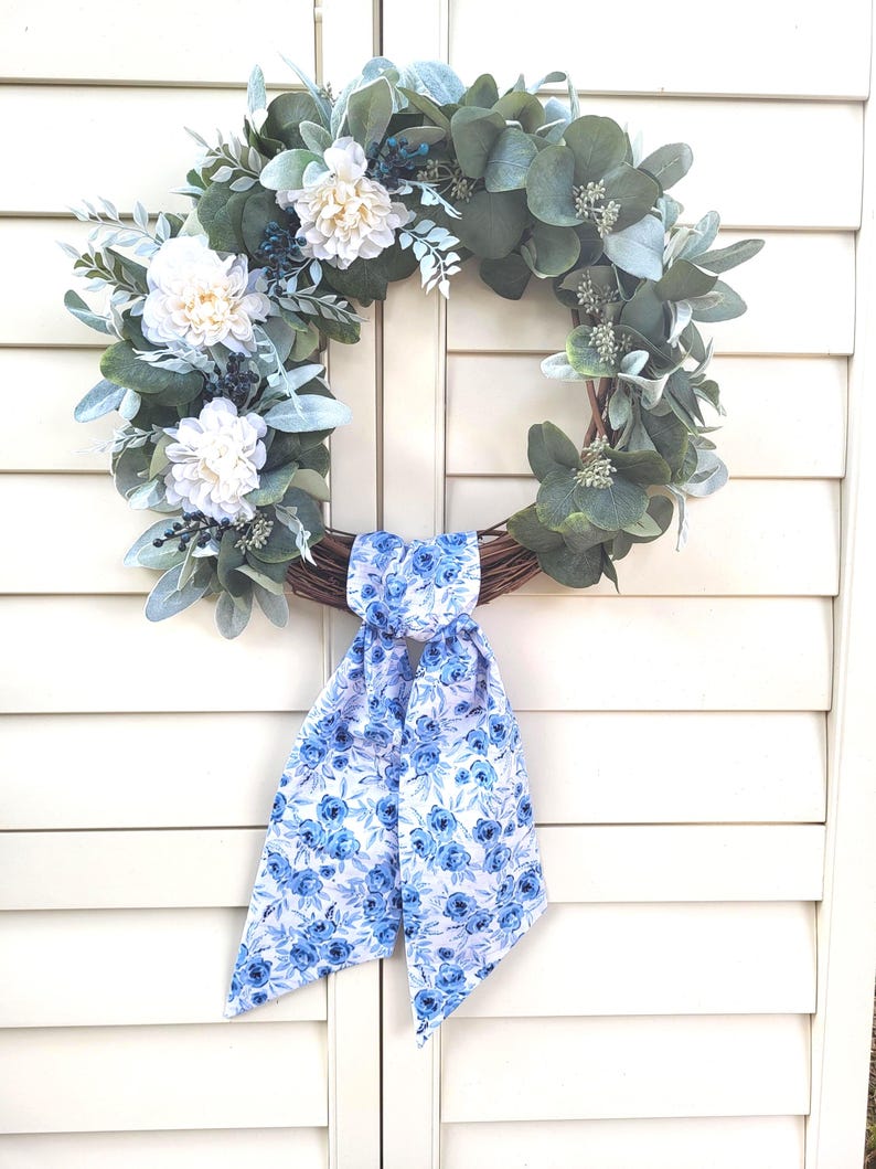 May include: A circular wreath constructed from brown twigs, adorned with green eucalyptus leaves, dusty miller foliage, and white dahlia flowers. A long fabric ribbon featuring a blue and white floral pattern is tied at the bottom of the wreath.