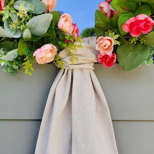 May include: A decorative wreath featuring artificial pink and peach flowers, green foliage, and dusty miller leaves. A long, beige fabric sash is tied in a knot at the center, with the fabric draping downward in vertical folds against a gray panel.
