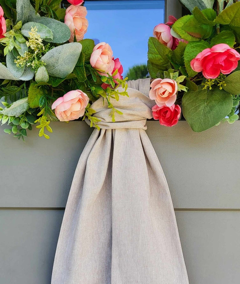 May include: A decorative wreath featuring artificial pink and peach flowers, green foliage, and dusty miller leaves. A long, beige fabric sash is tied in a knot at the center, with the fabric draping downward in vertical folds against a gray panel.