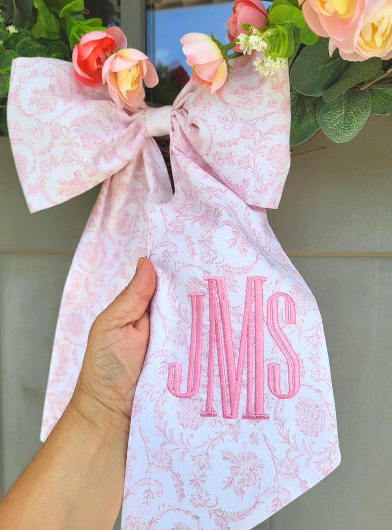 May include: A large fabric bow featuring a white and light pink floral toile pattern with long tails. The bow is accented with pink artificial flowers and greenery at the top. One tail is embroidered with the pink monogram letters JMS in a serif font.