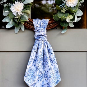 May include: A decorative wreath featuring green eucalyptus leaves and white flowers, accented with a white fabric scarf tied in a knot. The scarf displays a detailed blue floral toile pattern across its surface, hanging vertically from the center.