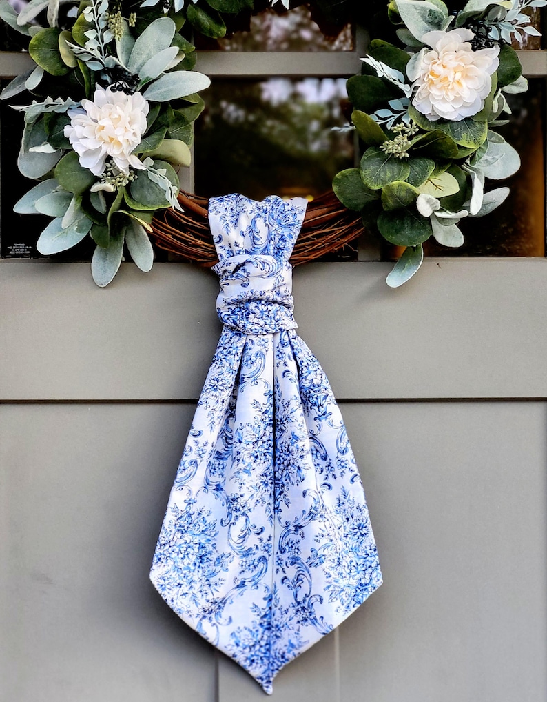 May include: A decorative wreath featuring green eucalyptus leaves and white flowers, accented with a white fabric scarf tied in a knot. The scarf displays a detailed blue floral toile pattern across its surface, hanging vertically from the center.