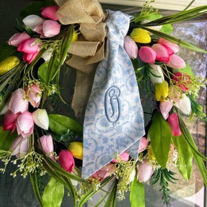 May include: A floral wreath featuring pink, white, and yellow tulips with green foliage, accented by a tan burlap bow and a light blue fabric ribbon with a white floral pattern and a dark blue embroidered letter G monogram in the center.