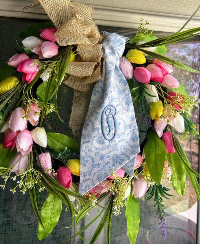 May include: A floral wreath featuring pink, white, and yellow tulips with green foliage, accented by a tan burlap bow and a light blue fabric ribbon with a white floral pattern and a dark blue embroidered letter G monogram in the center.