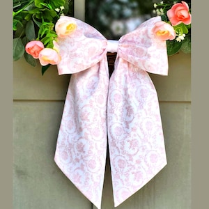 May include: A large decorative bow made of white fabric with a repeating light pink floral and scroll pattern. The bow features two long, pointed tails and is accented by small, soft pink artificial flowers tucked behind the top loops.