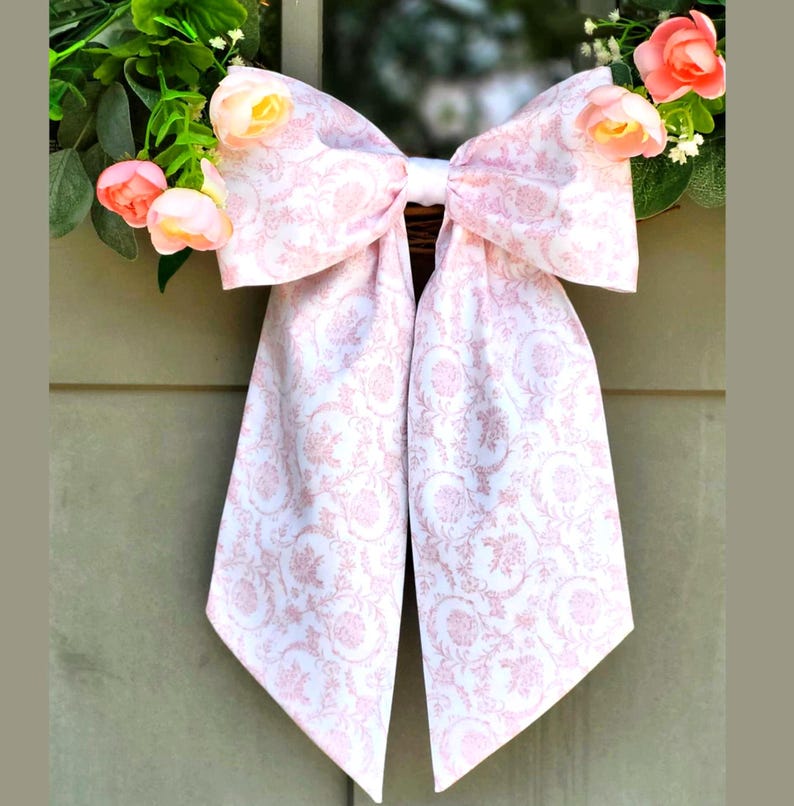 May include: A large decorative bow made of white fabric with a repeating light pink floral and scroll pattern. The bow features two long, pointed tails and is accented by small, soft pink artificial flowers tucked behind the top loops.