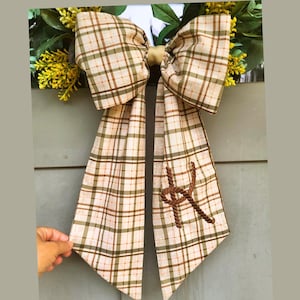 May include: A decorative bow featuring a beige, olive green, and brown plaid pattern with long, flowing tails. The right tail displays a brown, rope-style embroidered letter K. The bow is attached to a wreath with green leaves and yellow flowers.