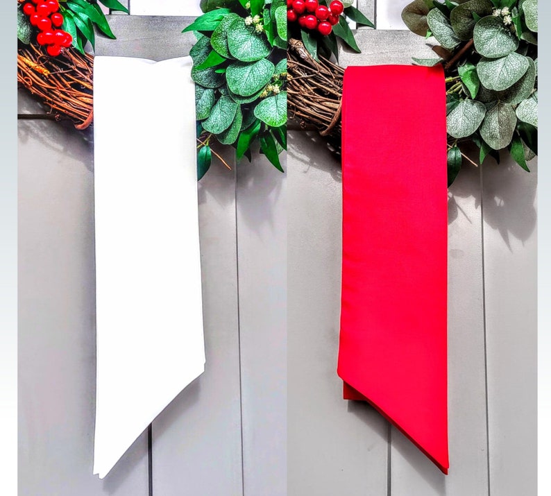 May include: Two long, rectangular ribbons, one white and one red, hang vertically against a gray background. The ribbons are positioned in front of a decorative wreath with green leaves and red berries.