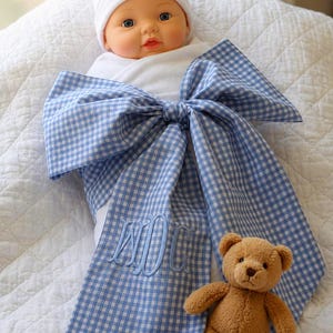 May include: A baby doll wearing a white beanie and white onesie, wrapped in a blue and white gingham blanket tied with a large bow. The blanket features an embroidered monogram with the letters MDC. A small brown plush teddy bear sits nearby.