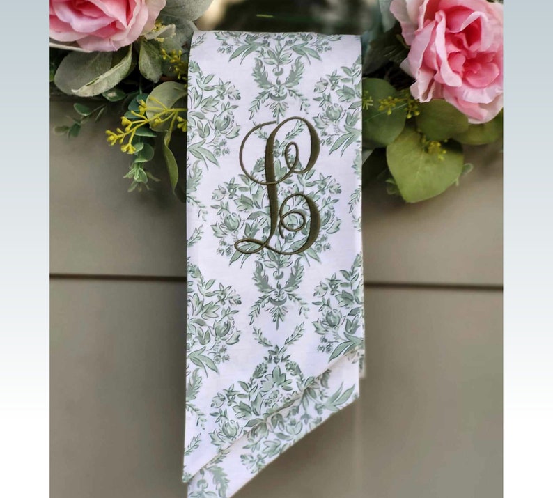 Pu&ograve; includere: Un nastro di tessuto bianco con un motivo botanico verde e una grande lettera B ricamata in verde scuro in corsivo. Il nastro &egrave; drappeggiato su una ghirlanda decorativa adornata con rose rosa e fogliame verde.