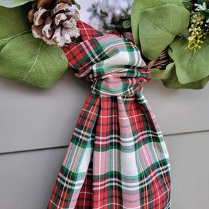 May include: A festive wreath featuring green eucalyptus leaves, a pinecone, and a red, green, and white plaid fabric bow. The wreath is designed for seasonal home decor.