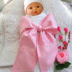 May include: A baby doll wearing a white beanie and a white swaddle blanket wrapped in a large pink and white gingham bow. The bow features an embroidered monogram in pink thread on the right side, with a soft texture and a classic fabric finish.