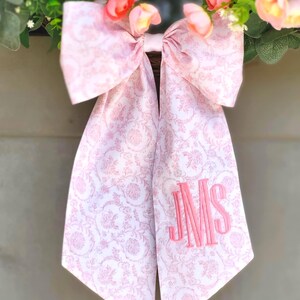 May include: A large decorative bow made of white fabric with a light pink floral toile pattern. The bow features two long tails, one of which is embroidered with the pink monogram letters JMS. It is attached to a wreath with pink flowers.