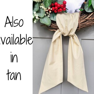 May include: A grapevine wreath adorned with faux greenery, red berries, and a tan fabric bow. The text "Also available in tan" is visible on the left side of the image. This decorative wreath is suitable for seasonal home decor.