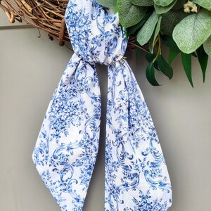 May include: A long, narrow fabric scarf featuring a white base with a repeating blue floral and scrollwork pattern. The scarf is tied in a simple knot, with two long, pointed ends hanging down, displaying the intricate botanical and ornate design.