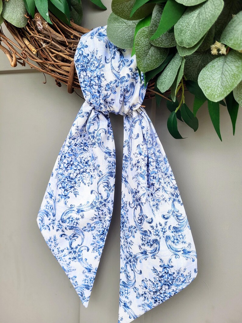 May include: A long, narrow fabric scarf featuring a white base with a repeating blue floral and scrollwork pattern. The scarf is tied in a simple knot, with two long, pointed ends hanging down, displaying the intricate botanical and ornate design.