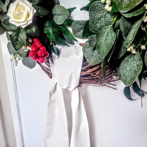 May include: A decorative wreath featuring a white rose, red berries, and various green leaves. The wreath is tied with a cream-colored satin ribbon. The base of the wreath appears to be made of twigs and branches.