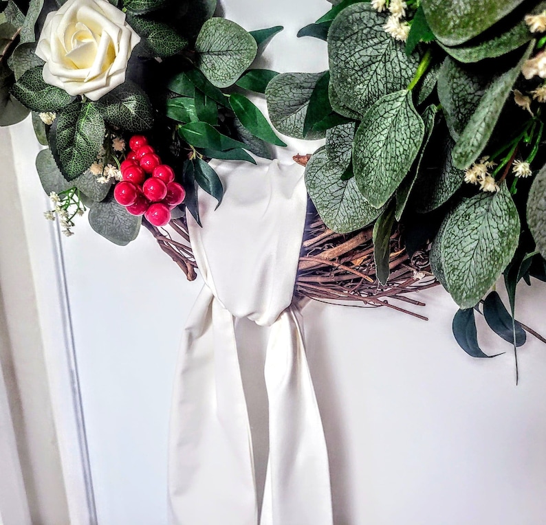 May include: A decorative wreath featuring a white rose, red berries, and various green leaves. The wreath is tied with a cream-colored satin ribbon. The base of the wreath appears to be made of twigs and branches.