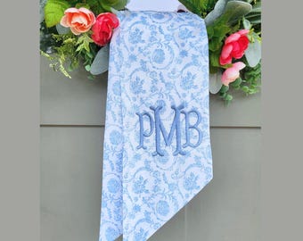 May include: A white fabric ribbon with a light blue floral pattern and a centered blue embroidered monogram featuring the letters PMB. The ribbon is draped over a decorative wreath adorned with green leaves and pink and orange flowers.
