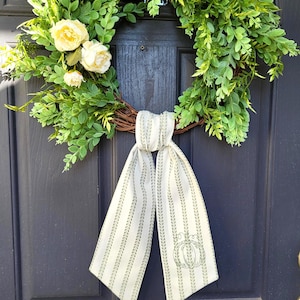 May include: A circular wreath constructed from brown twigs, adorned with lush green foliage and three pale yellow flowers. A long, off-white fabric ribbon with vertical green leaf-patterned stripes and a line-art pumpkin graphic hangs from the bottom.