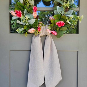 May include: A circular floral wreath featuring pink and peach-colored flowers, green foliage, and silver-green lamb's ear leaves. A long, plain beige fabric ribbon is tied in a knot at the bottom, with two long tails hanging down from the wreath.