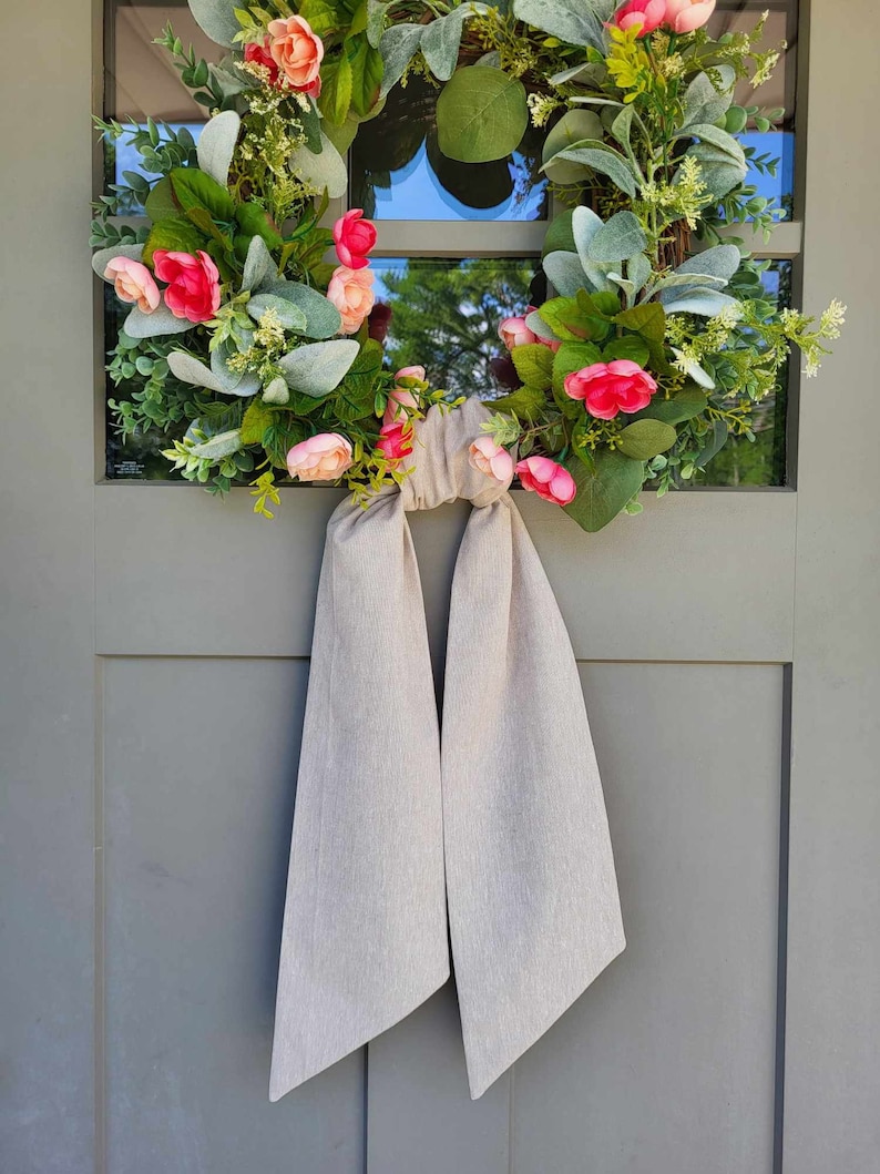 May include: A circular floral wreath featuring pink and peach-colored flowers, green foliage, and silver-green lamb's ear leaves. A long, plain beige fabric ribbon is tied in a knot at the bottom, with two long tails hanging down from the wreath.