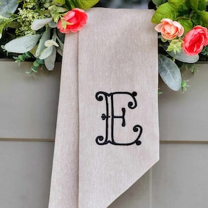 May include: A beige fabric ribbon featuring a black embroidered letter E in a decorative script font. The ribbon is draped behind a wreath adorned with artificial green leaves and small pink and peach-colored flowers.