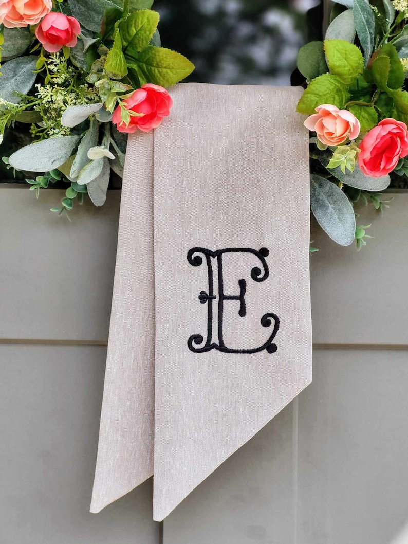 May include: A beige fabric ribbon featuring a black embroidered letter E in a decorative script font. The ribbon is draped behind a wreath adorned with artificial green leaves and small pink and peach-colored flowers.