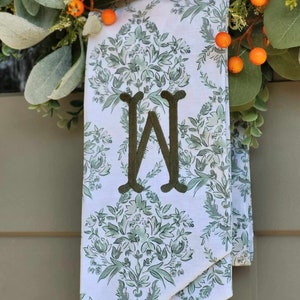 Pu&ograve; includere: Un drappo in tessuto bianco con un motivo floreale damascato verde e una grande lettera W ricamata in verde scuro al centro. Il tessuto &egrave; drappeggiato dietro una ghirlanda decorativa con foglie verdi e piccole bacche arancioni.