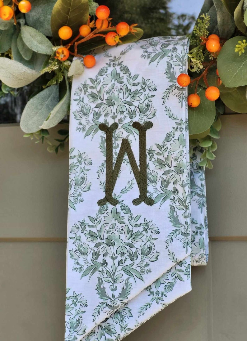Pu&ograve; includere: Un drappo in tessuto bianco con un motivo floreale damascato verde e una grande lettera W ricamata in verde scuro al centro. Il tessuto &egrave; drappeggiato dietro una ghirlanda decorativa con foglie verdi e piccole bacche arancioni.
