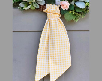 May include: A decorative wreath featuring a brown twig base adorned with green eucalyptus leaves and soft pink flowers. A long, yellow and white gingham fabric ribbon is tied in a knot at the top, with two long tails hanging down the center.