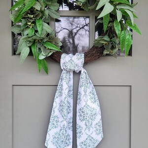 Pu&ograve; includere: Una ghirlanda circolare realizzata con ramoscelli marroni, decorata con foglie verdi rigogliose e piccoli boccioli gialli. Un lungo nastro di tessuto bianco con un motivo floreale damascato verde salvia &egrave; annodato alla base della ghirlanda.