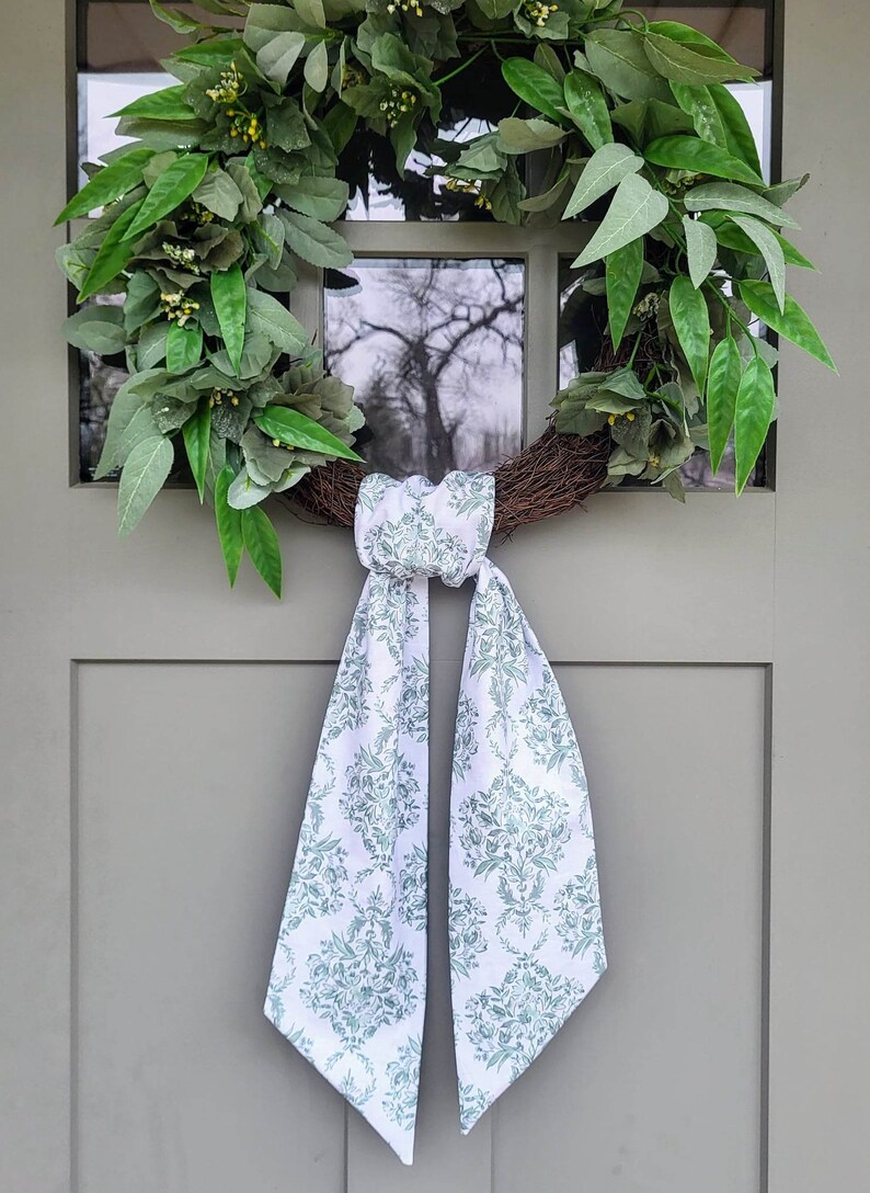 Pu&ograve; includere: Una ghirlanda circolare realizzata con ramoscelli marroni, decorata con foglie verdi rigogliose e piccoli boccioli gialli. Un lungo nastro di tessuto bianco con un motivo floreale damascato verde salvia &egrave; annodato alla base della ghirlanda.