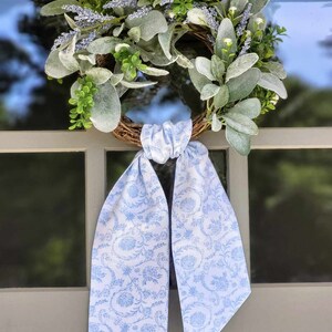 May include: A round wreath featuring a brown twig base adorned with fuzzy sage green leaves, small white flowers, and sprigs of lavender. A long, light blue fabric ribbon with a white floral pattern is tied in a knot at the bottom of the wreath.