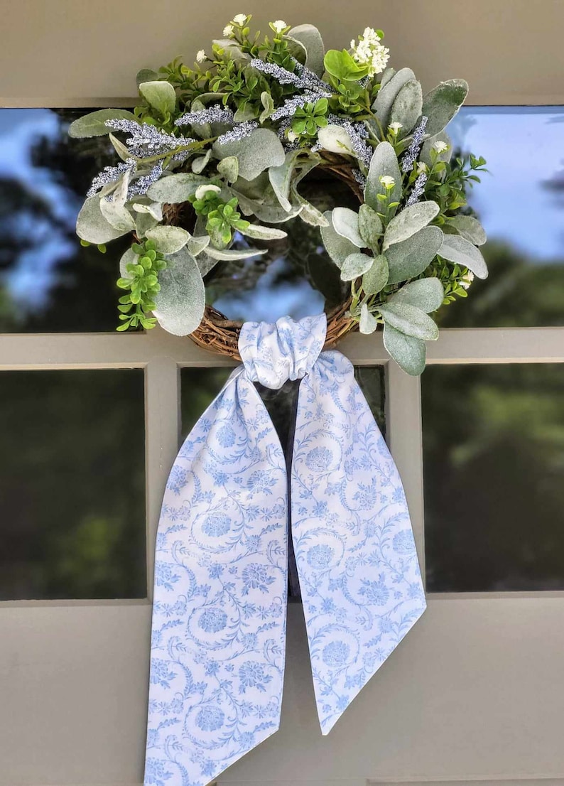 May include: A round wreath featuring a brown twig base adorned with fuzzy sage green leaves, small white flowers, and sprigs of lavender. A long, light blue fabric ribbon with a white floral pattern is tied in a knot at the bottom of the wreath.