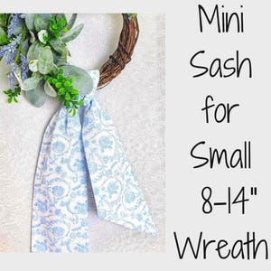 May include: A light blue and white floral patterned fabric sash tied to a small brown twig wreath decorated with green leaves and purple lavender sprigs. The text reads: Mini Sash for Small 8-14" Wreath. The design features a repeating botanical print.