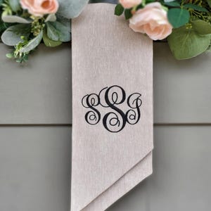 May include: A rectangular beige fabric banner featuring a black embroidered monogram with three interlocking script letters. The banner hangs vertically and is accented by two clusters of artificial light pink roses and green eucalyptus leaves.
