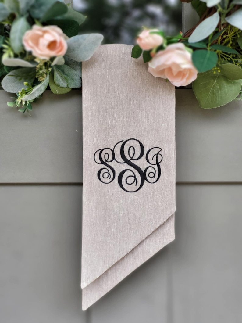 May include: A rectangular beige fabric banner featuring a black embroidered monogram with three interlocking script letters. The banner hangs vertically and is accented by two clusters of artificial light pink roses and green eucalyptus leaves.