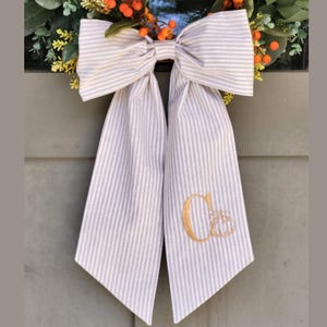 May include: A large decorative bow made of light gray and white striped fabric, featuring two long hanging tails. The right tail is embroidered with a gold-colored letter C and a small pumpkin icon, positioned beneath a wreath of green leaves and orange berries.